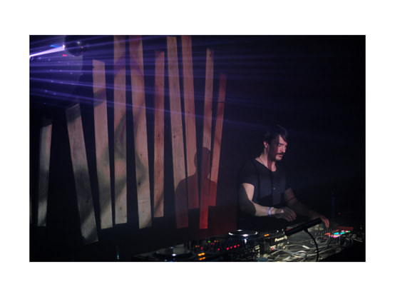 handsome young man standing in front of wooden panels with viduals projected on, playing electronic music instruments live