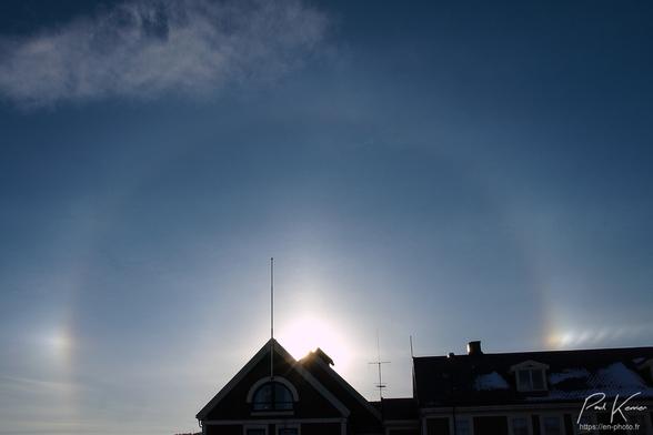 Photo en couleur présentant un petit halo de 22° autour du soleil avec ses 2 parhélies (sundogs en anglais) bien visibles et colorés, prolongés de chaque côté par le début du cercle parhélique également bien coloré. La plus grande partie du soleil est cachée derrière le faîte du toit d'un bâtiment afin de modérer la surexposition. Le ciel est pratiquement bleu à l'exception d'un Léger nuage dans le coin supérieur gauche de la photo, le soleil et son halo étant pratiquement centrés.