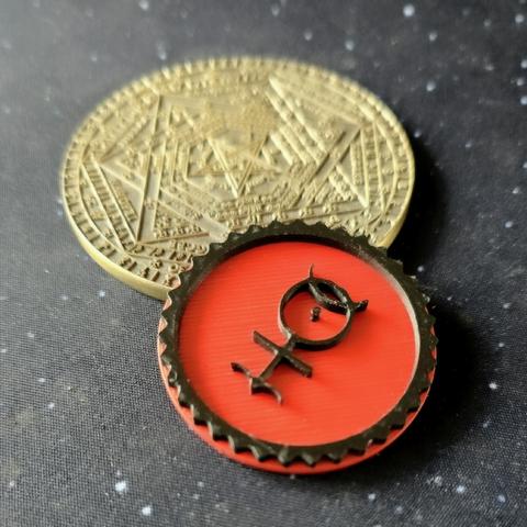 A detailed metal coin with intricate geometric and alchemical engravings lies on a dark, star-speckled surface. In the foreground, a red and black token with a serrated edge features a raised symbol resembling the astrological sign for Mercury, stylized with small horns and additional embellishments.
