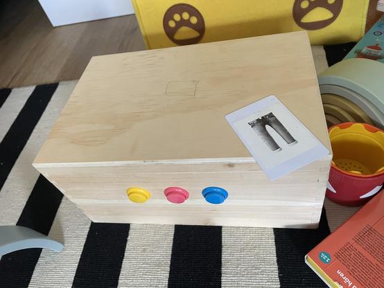 A wooden box with three colored buttons (yellow, pink, and blue) on one side. A rfid card depicting a pair of pants rests on top. The background includes a colorful toy and books on a striped rug.