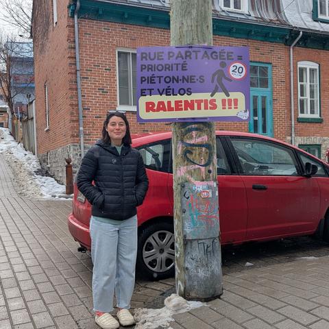 Une personne souriante, les mains dans les poches, est debout dans une  rue pavée, à côté d'un poteau de bois. Une affiche mauve est installée sur le poteau. Il y est écrit en noir : "rue partagée = priorité piéton·ne·es et vélos" , et en rouge sur fond blanc , en plus gros caractères ; " RALENTIS" . Une icône de piéton figure aussi sur l'affiche, ainsi que "20 km/h" dans un cercle blanc à la bordure rouge.  En arrière-plan, une maison de brique au toit de fer et au solage de pierres, typique du quartier St-Jean-Baptiste à Québec. Une voiture rouge est stationnée trop près de l'intersection.