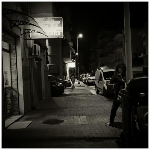 A woman waits as she checks her mobile phone outside a pizzeria on an urban pavement illuminated by the sign. The faint light of the mobile phone barely illuminates her face. In the background illuminated by a street lamp, a person is walking apparently in a hurry. At the side of the pavement a row of parked cars