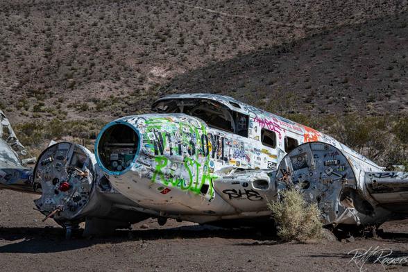 Photograph of the abandoned fuselage of an old prop plane, stripped of parts and engine, lying on the desert floor.