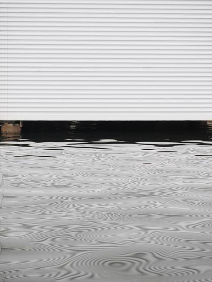 The image shows a scene with water partially covering an area in front of what appears to be a white, corrugated metal door or wall. The water has a reflective surface with a wavy, textured appearance.