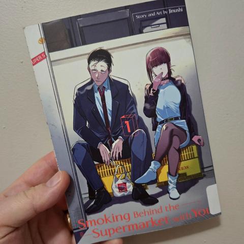 Photograph of a white person's hand holding "Smoking Behind the Supermarket With You," volume 1. Image shows an older man in a suit next to a younger woman with purple hair in a skirt and leather jacket smoking. They are sitting on crates against a wall.