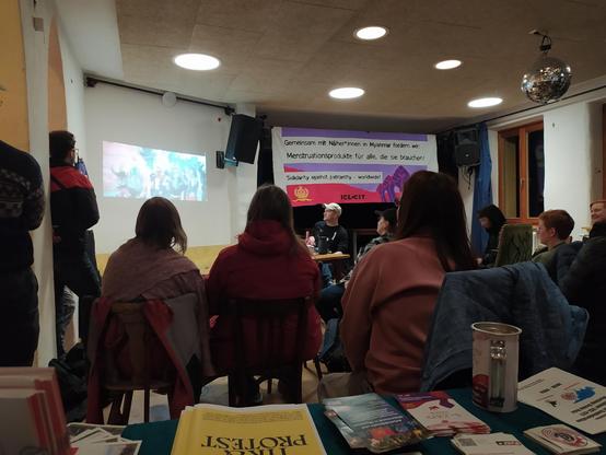 Im Hintergrund hängt ein Demo Banner mit der Schrift: Gemeinsam mit Näher*innen in Myamar fordern wir Menstruationsprodukte für alle, die sie brauchen! Solidarity against patriachy worldwide. Im Vodergrund sieht man den Rücken mehrerer sitzenden Personen, die auf eine Projektion an der Wand schauen, eine Person erzählt etwas zu diesem Bild