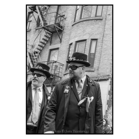 (English follows) Photographie noir et blanc, format portrait, ratio 2x3. L'image est prise en contre-plongée. Elle saisit au premier plan deux marguillers, portant des lunettes noires et des chapeaux décorés de trèfles ; épinglées sur leur veste, diverses décorations soulignent leur appartenance à la communauté irlandaise. Les deux hommes regardent vers le public, dans la même direction. À l'arrière plan, se dresse un immeuble typique de Montréal : revêtement de briques, escaliers de secours métalliques. 

Black and white photograph, portrait format, 2x3 ratio. The image is taken from a low angle. In the foreground are two marshals, wearing dark glasses and hats decorated with shamrocks; pinned to their jackets are various decorations emphasizing their membership of the Irish community. Both men are looking in the same direction towards the audience. In the background stands a typical Montreal building: brick cladding, metal fire escapes.