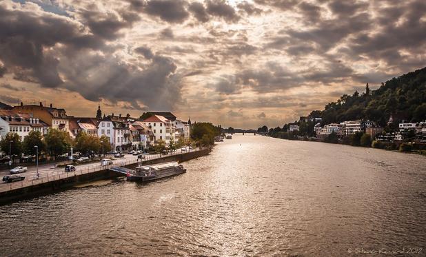 A view from a bridge looking down a river as it stretches up the frame, with the bank on either side. On the left are classic Germany buildings with the street running alongside the river and a few cars parked and driving along. There is a long docked barge with the gangway from it to the quayside. On the right bank are some buildings and a tree covered hill. The sky is dramatic with displays of clouds and a pink tone from the setting sun which is reflected in the water Another bridge can just be seen in the far distance.