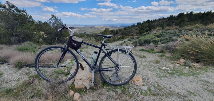 My trusty old Trek hybrid bicycle parked on a rocky dirt path in a rural landscape. The background features rolling hills, green shrubs, and a distant mountain range under a partly cloudy blue sky. The terrain consists of dry grass, small rocks, and patches of greenery.