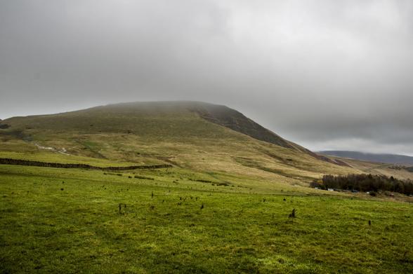A dark green landscape rising to the peak of Mam Tor, shrouded in rainclouds.