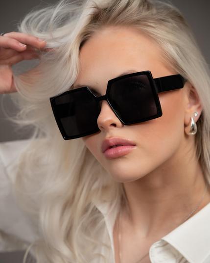 The image shows a person wearing large, square-shaped sunglasses. They have light blonde hair and are wearing a light-colored shirt. The person is also wearing earrings and a necklace, giving a stylish and fashionable appearance.