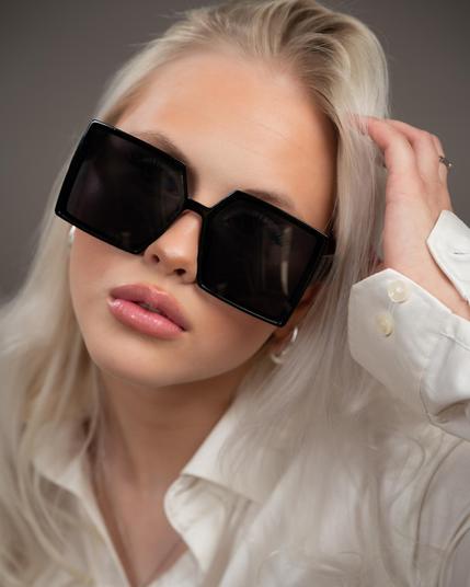 The image shows a person with long, platinum blonde hair wearing large, black, square sunglasses. They are dressed in a white shirt, and their hand is touching their hair. The lighting highlights their facial features, and their expression is neutral or slightly pouty.
