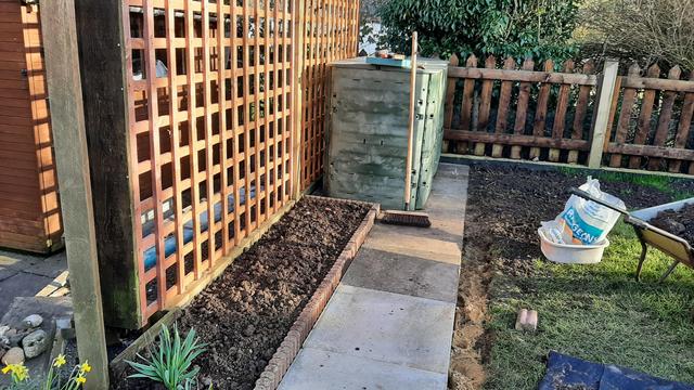 Golden brown squared garden trellis on the left with new flower bed to its right. Green compost bin at the rear. Grass on the right of new paving in centre.