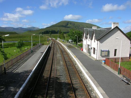 Achnasheen. The image shows a view from a railway bridge of a railway extending away from the camera. It is twin track in the foreground but becomes single track in the middle distance, and then curves to the left. In the foreground is a station with platforms either side. On the right is a white building with a slate roof that fronts on to the station platform. The scene is in sunlight.