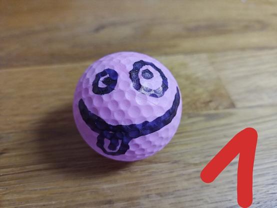 Wir sehen einen pink farbenen Golfball. Auf ihm ist eine Gesicht aufgemalt mit großen Runden Augen und einem breiten Grinsemund, der uns die Zunge raus streckt