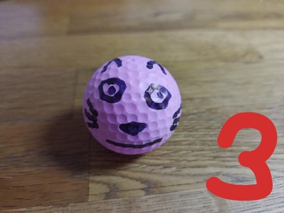 vor uns liegt ein pink farbener Golfball, auf dem ein Katzengesicht aufgemalt ist.
Die Katze hat große runde Augen und lächelt mit schmalen Lippen