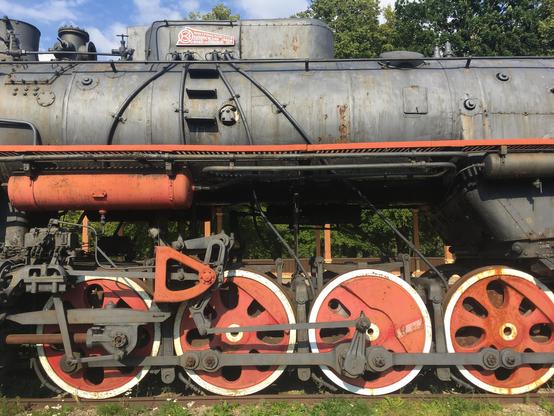 A partial side shot of the center of a steam locomotive. Most of the top barrel is plain metal, slightly rusted. We also see red painter metal, most notably in the four “wheels” and some smaller red barrel on top (I know nothing about train terminology, sorry). The red wheels have white rims and are connected with a big metal rod.