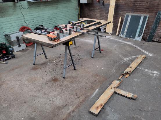 Werkplaats met een poort op schragen en lijmklemmen om de poort uit elkaar te drukken zodat deze gerestaureerd kan worden. Op de grond een gebroken houten stijl.