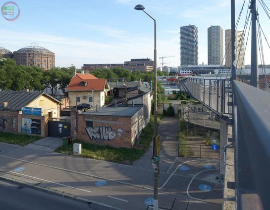 Straße mit Gewerbebau, dahinter ein Wohngebäude mit Wanduhr, an der ein darübersituierter vom WInd gesteuerter Fisch hängt (Kunstwerk "Winduhr oder Das Leben ein Hauch" von Alfredo Barsuglia).