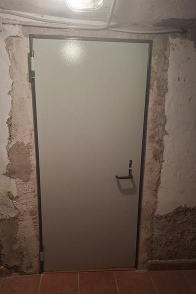 The image shows a metal door with a light-colored, possibly off-white or gray, textured surface. The door has a dark metal frame and a black handle with a keyhole. The surrounding walls appear to be unfinished or partially renovated, with patches of exposed rough plaster and some areas of smooth white coating. The floor is covered with reddish-brown tiles, and there is a light fixture above the door with electrical conduits running along the ceiling. The overall setting suggests that this door is installed in a basement or utility room.