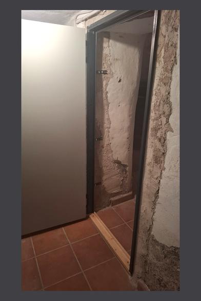 This image shows an unfinished doorway installation in a room with rustic, partially renovated walls. A dark metal door frame is being installed, with metal brackets protruding from its sides, likely for securing it to the wall. The lower part of the frame is supported by a wooden board, which helps with alignment and leveling.
In the background, another closed door is visible, which appears older and has a worn surface with some scratches or stains.
The walls have a mix of exposed plaster and rough, unfinished patches, suggesting that renovation work is ongoing. The floor is covered with reddish-brown terracotta tiles.
