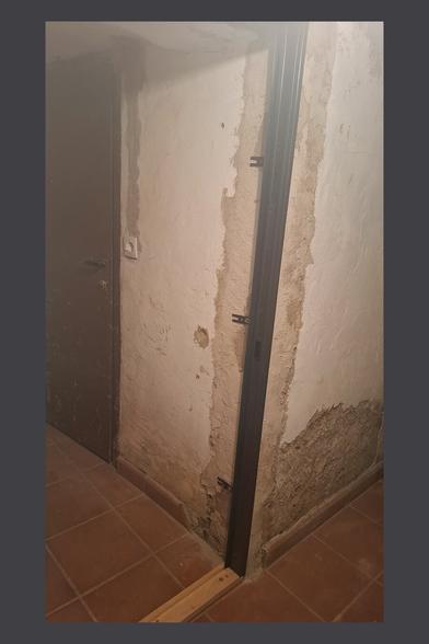 This image shows an unfinished doorway installation in a room with rustic, partially renovated walls. A dark metal door frame is being installed, with metal brackets protruding from its sides, likely for securing it to the wall. The lower part of the frame is supported by a wooden board, which helps with alignment and leveling.

In the background, another closed door is visible, which appears older and has a worn surface with some scratches or stains. A light switch is mounted on the wall near this door.
The walls have a mix of exposed plaster and rough, unfinished patches, suggesting that renovation work is ongoing. The floor is covered with reddish-brown tiles