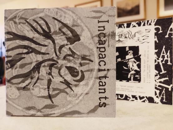 Fold out cd cover. Mostly gray, abstract art, with "Incapacitants" written sideways down the right side. Part of the inside is visible, where there is a copy of one of the original tape artworks. A black and white photo of two men in some fighting match is seen.