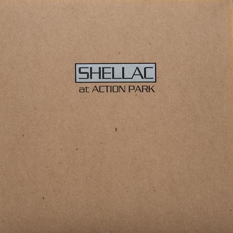 The album art for At Action Park by Shellac. It looks like matte or satin unprinted card stock or light kraft paper, with a small logo in the upper half, centred horizontally. The words are printed in black in a boxy typeface with thin strokes and right angles. The word SHELLAC is in a black outlined silver rectangle.