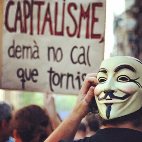 A banner with the message "Capitalism, you don't need to come back tomorrow" in catalan on the left. On the right someone with a V for Vendetta mask.