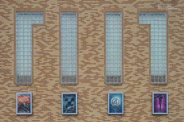 A tan and brown brick wall has four glass block windows.  Under each window is a movie poster.