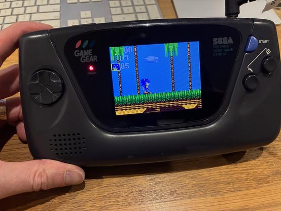 A black Sega GameGear handheld console with a large color screen showing Sonic the Hedgehog.