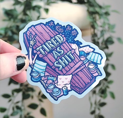 hand holding up a sticker of a wooden coffin with a skeleton hand sticking out throwing a peace sign, titled "tired as shit"