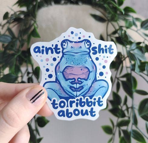hand holding up a sticker of a frog sitting like a human, titled "ain't sh*t to ribbit about"
