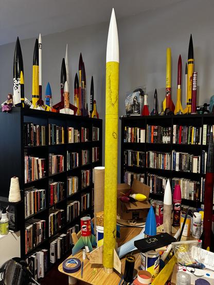 A tall model rocket covered in yellow fiberglass, in front of bookshelves, and surrounded by other rocketry projects.
