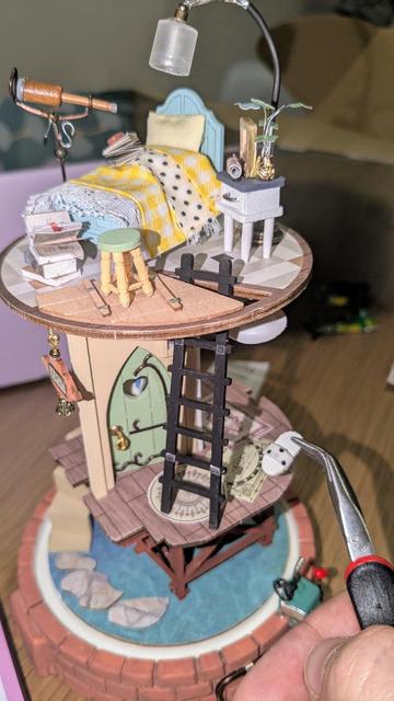 Torre semimontada, se ve mi mano con unas pinzas sosteniendo unas mini zapatillas de estar por casa blancas y con puntos negros, hechas de tela y cartón.

la torre tiene forma de cilindro sin paredes, el piso de arriba ya tiene la cama, el telescopio y la mesilla, hay una escalera de mano para subir, en el piso bajo está montada una puerta y alfombras