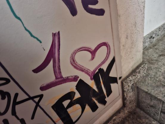 Ein lilafarbenes "1💜" an eine Wand mit weiteren Grafittis gesprüht. Unten rechts sind ein paar graue Treppenstufen.