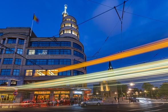 Look up, wide angle shot at slow shutter speed of Café Belga and Flagey art deco building on Place Flagey in Brussels at the evening's blue hour. The café is filled with customers and bathed in red light and the light trails of a tram passing by in the foreground complete the composition, adding a nice color contrast with a bright orange line against the dark blue sky and a light green line toping the red glow of the café.