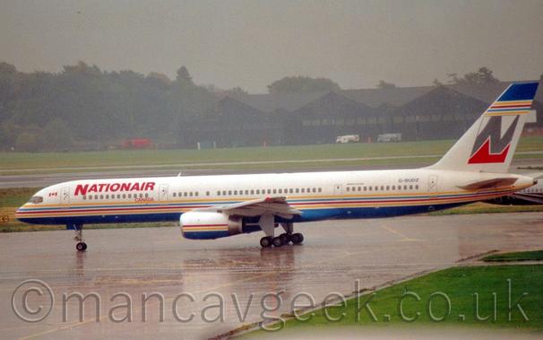 Side view of a white, twin engined jet airliner taxiing from right to left.
The plane has a light blue belly with thin yellow, blue, and red stripes running along the body, a design replicated on the engine pods and in reverse on the tip of the tail.
The tail has the thin grey outline of a square, containing a large grey upper case letter "N", with the inside of the square under the letter being red.
Saturated concrete apron fills most of the foreground, small splashes of raindrops can be seen on the ground, with a wide strip of grass on the right.
More grass in the background runs up to black hangars in the distance, with trees vanishing into rainy grey sky beyond that.