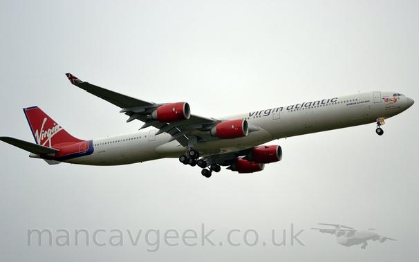 Side view of a white, 4 engined jet airliner flying from left to right at a low altitude, with undercarriage lowered and flaps extended from behind the wings, suggesting it is about to land.
The plane has purple "Virgin Atlantic" titles on the upper forward fuselage, with black "A340-600" titles a bit further forward and lower.
The engine pods mounted under the wings are red.
The tail and part of the rear fuselage are the same red, outlined in purple, with seemingly hand-written white "Virgin" titles.
There is an image of a woman wearing a red leotard and trailing a Union Flag from her arms in a flying pose under the cockpit windows.