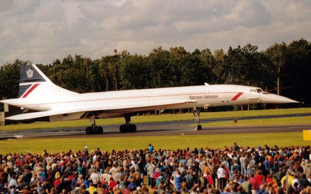 Side view of a long, thin, and pointy white, 4 engined jet airliner, taxiing from left to right along a black runway.
There is a thin, red stripe running along the body, with a red arrowhead just behind the cockpit.
There are black "British Airways" titles on the lower forward fuselage.
The top of the tail is dark blue, with a white crest, while the bottom half has a blue triangle and diagonal red stripe.
The registration "G-BOAB" is on the lower rear fuselage in black.
In the foreground, a large crowd is gathered, watching the plane, separated from it by a large area of grass.
More grass is visible in the background under the planes body, leading up to tall trees in the distance, under fluffy grey clouds.