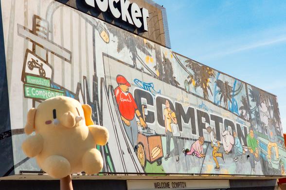 Photo of the Creature Mastodon stuffed toy in front of a giant outdoor mural in Compton during a bright, sunny day. The Creature's held in the air on the arm of a person offscreen, its back to the untitled mural about life in Compton and LA pride by Mel Depaz, AngelOnce, and GoopMassta at Foot Locker in Compton.