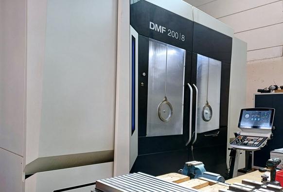 A DMF 200|8 CNC machine in an industrial workshop. The large machining center has closed double doors with viewing windows. A control panel with a screen and various tools are placed on a nearby workbench.