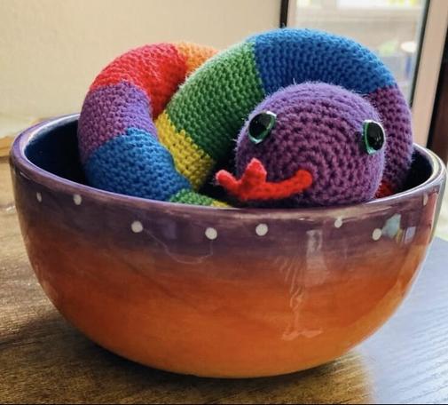 A crochet rainbow colored snake winding over itself inside of a sunset painted bowl. The snake is looking at you with green eyes, a purple head and red tongue sticking out. The bowl rests on a wood tabletop.