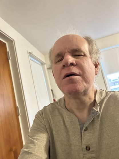 The image depicts an older man with white hair. He is wearing a light-colored shirt. He appears to be in a room with a wooden door and a window with blinds. The man is looking directly at the camera, his eyes closed, and his mouth slightly open, giving the impression of a contemplative or serene expression. The room seems to be well-lit, possibly by natural light coming through the window.