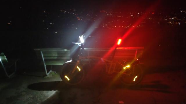 Mein Brompton Electric G-Line steht nachts in voller Beleuchtung an eine Parkbank gelehnt. Profilansicht. Das Bild hat heftige Lens Flares von unten links nach oben rechts. Von der Seite sieht man vor allem die beiden Knog Cobber Big Akkuleuchten an Lenker- und Sattelstange. Im Hintergrund die Lichter der Stadt.