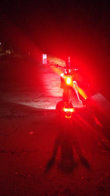 Mein Brompton Electric G-Line steht nachts in voller Beleuchtung an eine Parkbank gelehnt. Ansicht von hinten. Das Bild hat heftige Lens Flares von unten rechts nach oben links. Man sieht die Knog Cobber Big Akkuleuchte an der Sattelstange und das vom E-Bike-Akku gespeiste Rücklicht am Gepäckträger.