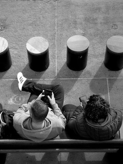 Black and white. View from above, looking over a wall at two young men sitting on a bench below, holding their phones, with a row of low, round, tube-shaped stools in front of them.