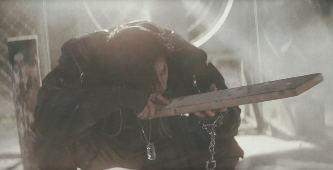 Bang Chan crouching down and using a plank of wood like something to sight down. He's wearing a dogtag pendant on a chain.