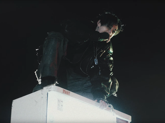 Bang Chan crouching on top of a vending machine. He's wearing a dogtag pendant on a chain.