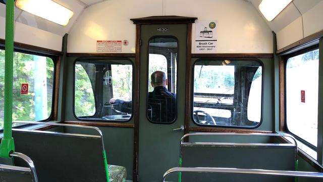 The image shows the interior of a vintage tram or trolley car, with a view towards the driver's area. The driver is seated, facing forward, and visible through a central glass door. The inside of the tram has green seats with metal frames and handrails. There are signs on the walls, including one mentioning the "Brush Car 80th Anniversary 1937 - 2017." The windows show a blurred outdoor scene, indicating movement. The lighting inside is provided by overhead fluorescent lights, and there are no-smoking signs on the walls.